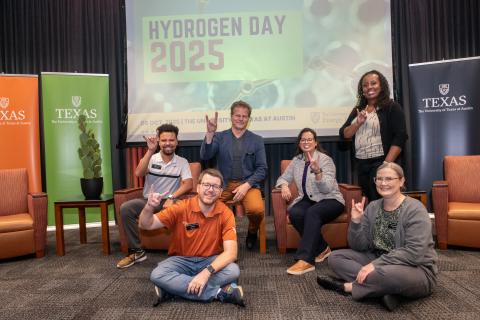 Six members of the EI staff are seated on a stage, giving the Hook 'Em hand sign
