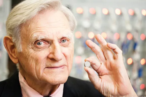 John Goodenough holds a coin cell battery between his index finger and thumb