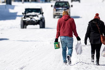 two people walk through snow carrying groceries as vehicles approach in the other direction